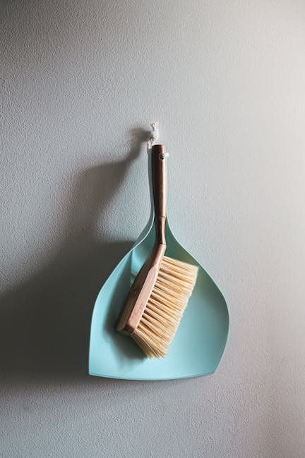 A light blue plastic dustpan hangs on a plain white wall, with a wooden-handled brush resting inside it. The brush has light-colored bristles, and both tools are positioned vertically. The scene sugge