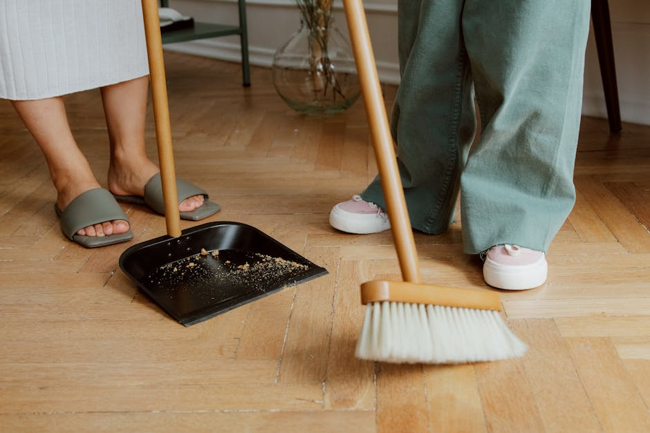 A light blue plastic dustpan hangs on a plain white wall, with a wooden-handled brush resting inside it. The brush has light-colored bristles, and both tools are positioned vertically. The scene sugge