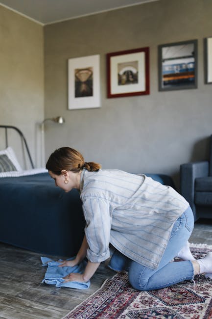A professional cleaner dressed in a white protective suit, gloves, and face mask is performing deep cleaning on a beige, textured carpet in a bright, modern living room. The cleaner is using a black h